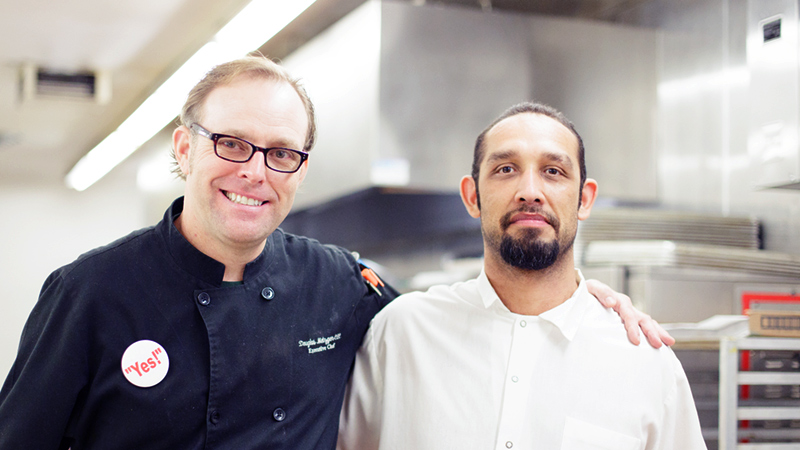 A chef and man posing together in commerical kitchen.