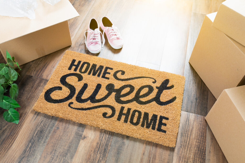 A welcome mat hat reads "home sweet home" rests on hardwood floor with boxes and a pair of sneakers nearby.