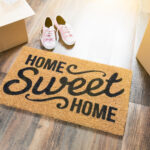 A welcome mat hat reads "home sweet home" rests on hardwood floor with boxes and a pair of sneakers nearby.