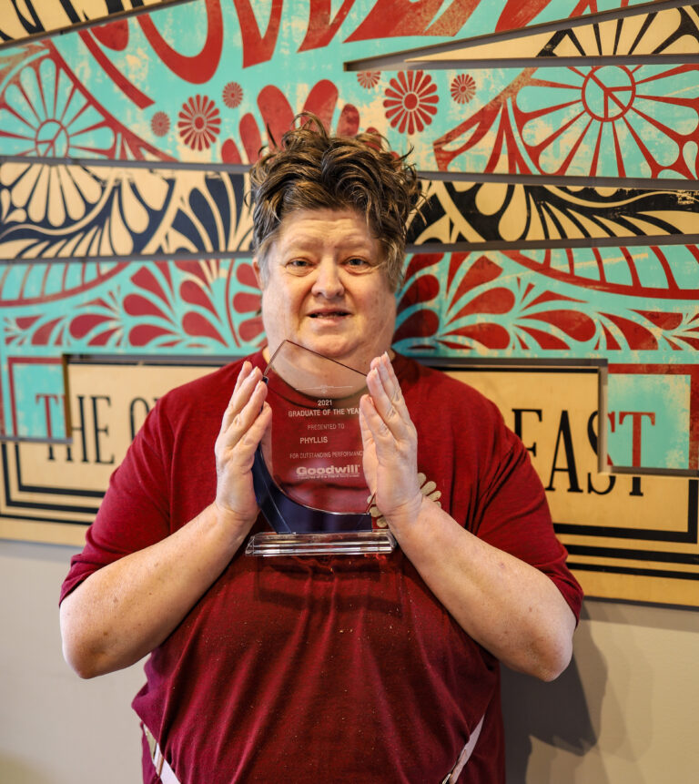 A woman in a red shirt stands in front of a patterned wall, holding a glass transparent award.