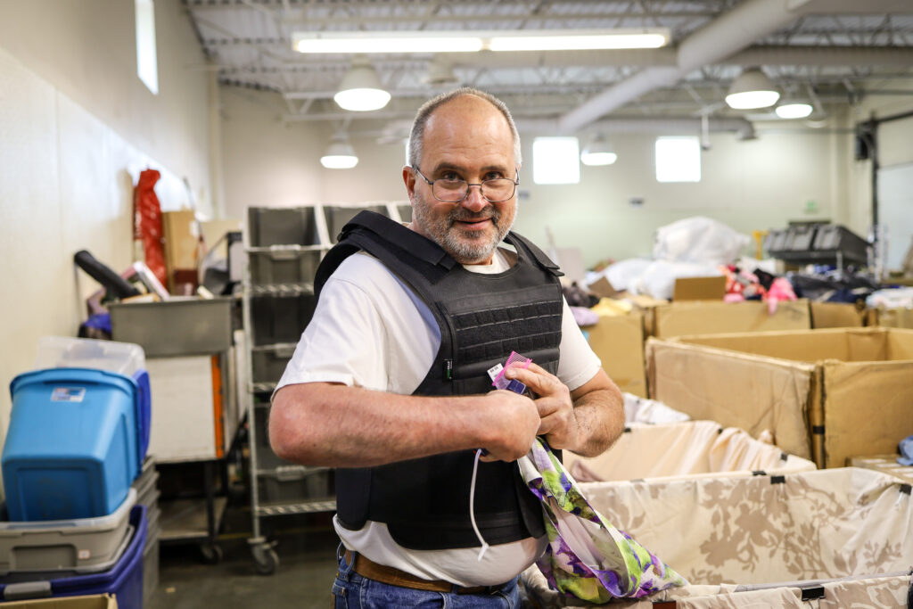 A man wearing a black protective vest is smiling at the camera while puncturing tags onto clothing at a textile sorting station.