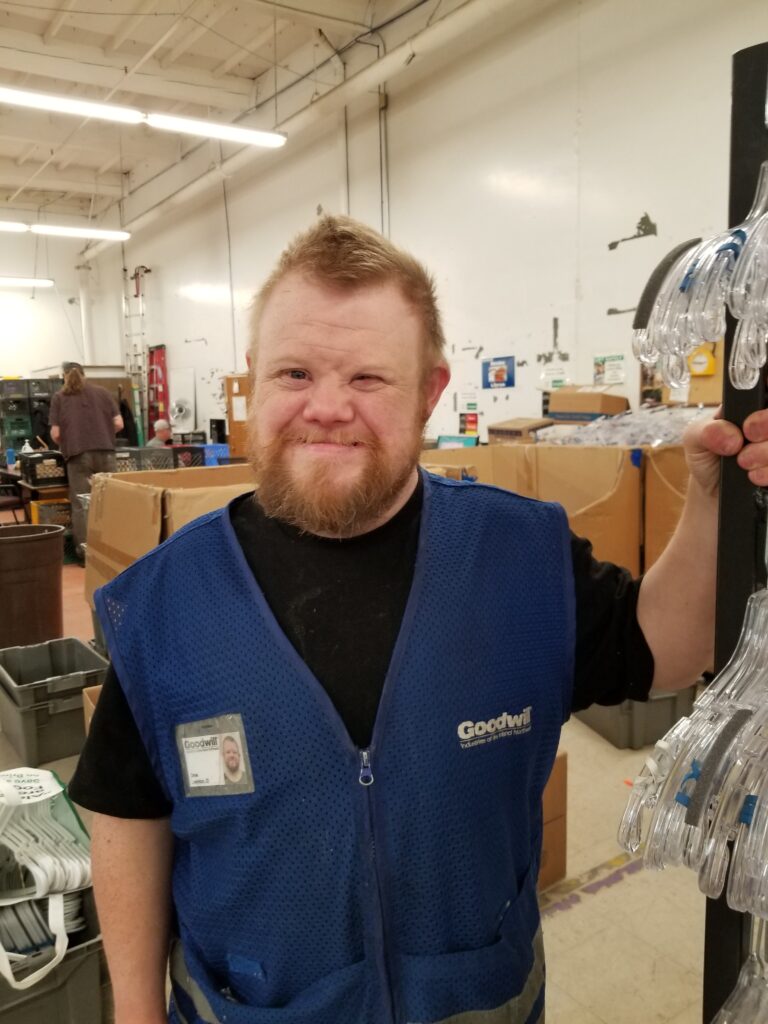 A man wearing a blue work vest with the word "Goodwill" on it, stands with his left hand gripping a textiles rack with empty hangers on it. He is smiling.