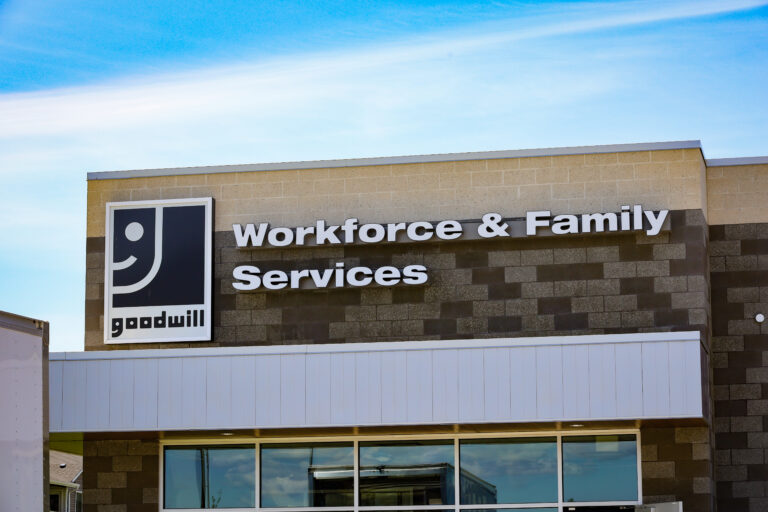 A building with the words "Workforce and Family Services" and a smiling "G" Goodwill logo on the top front. The building is against a light blue sky background.