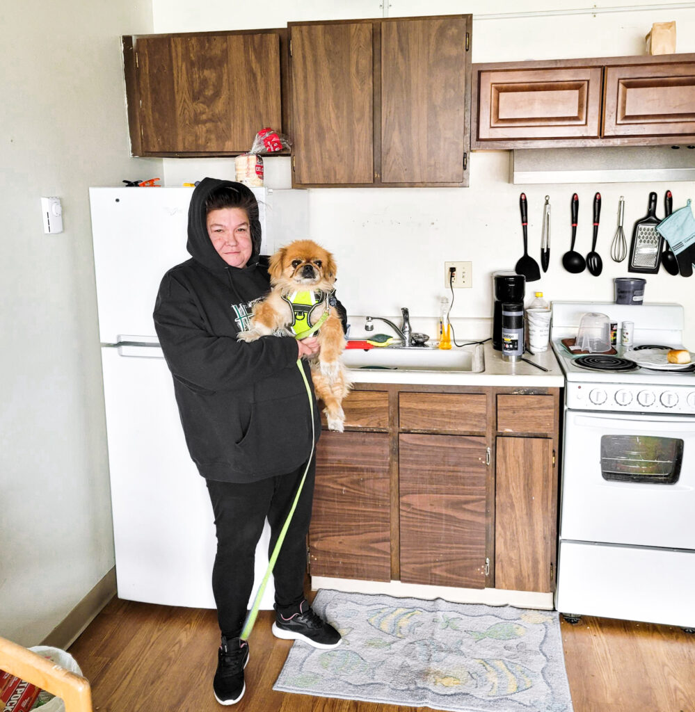 A woman holds a small dog and stands in a modest kitchen.