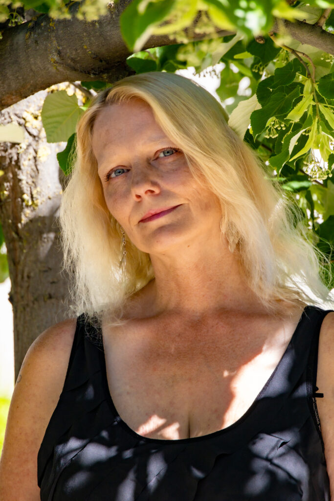 A woman with platinum blonde hair wears a black tank top stand under in the shade under a tree and smiles.