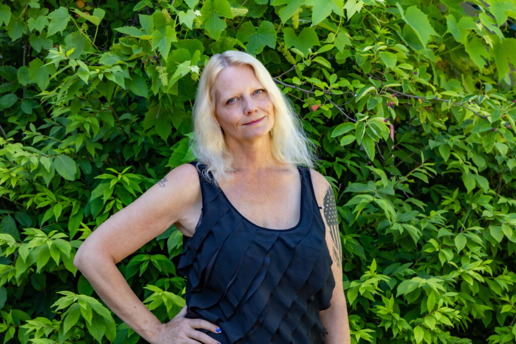 A woman with platinum blonde hair, wearing a black tank top shirt stands in front of a large green bush smiling with her right hand on her hip.