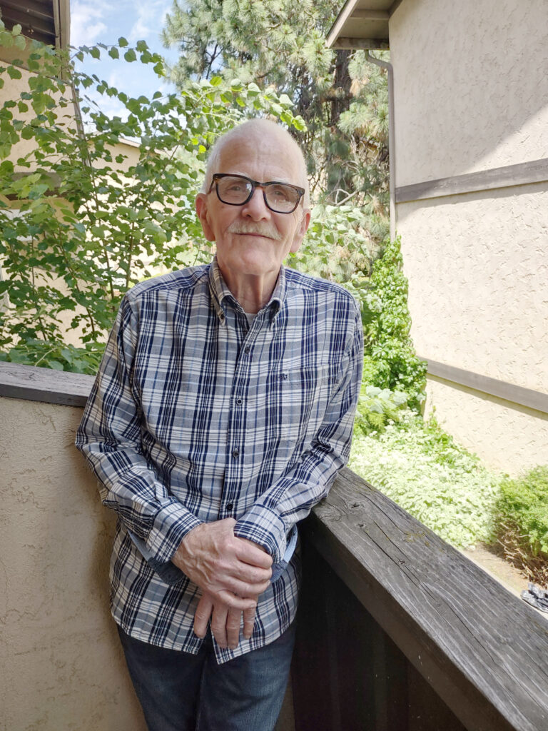 An elderly man wearing a long sleeved plaid button up shirt and thick dark rimmed glasses stands on an outdoor balcony and smiles at the camera.