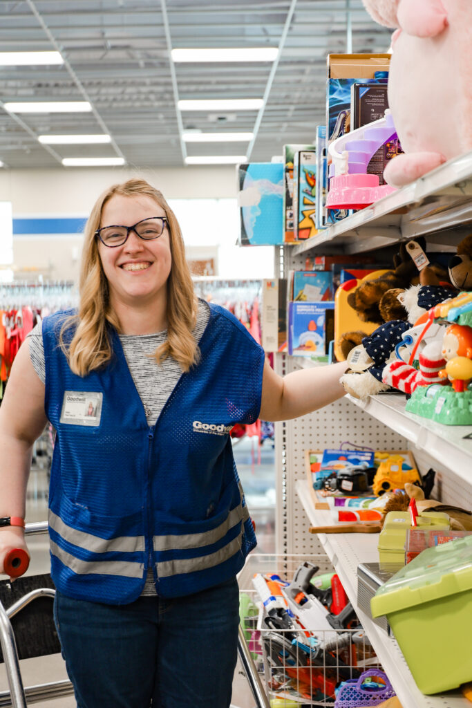 A woman wearing a blue Goodwill vest is standing next to a shelf of donated goods, smiling at the camera.