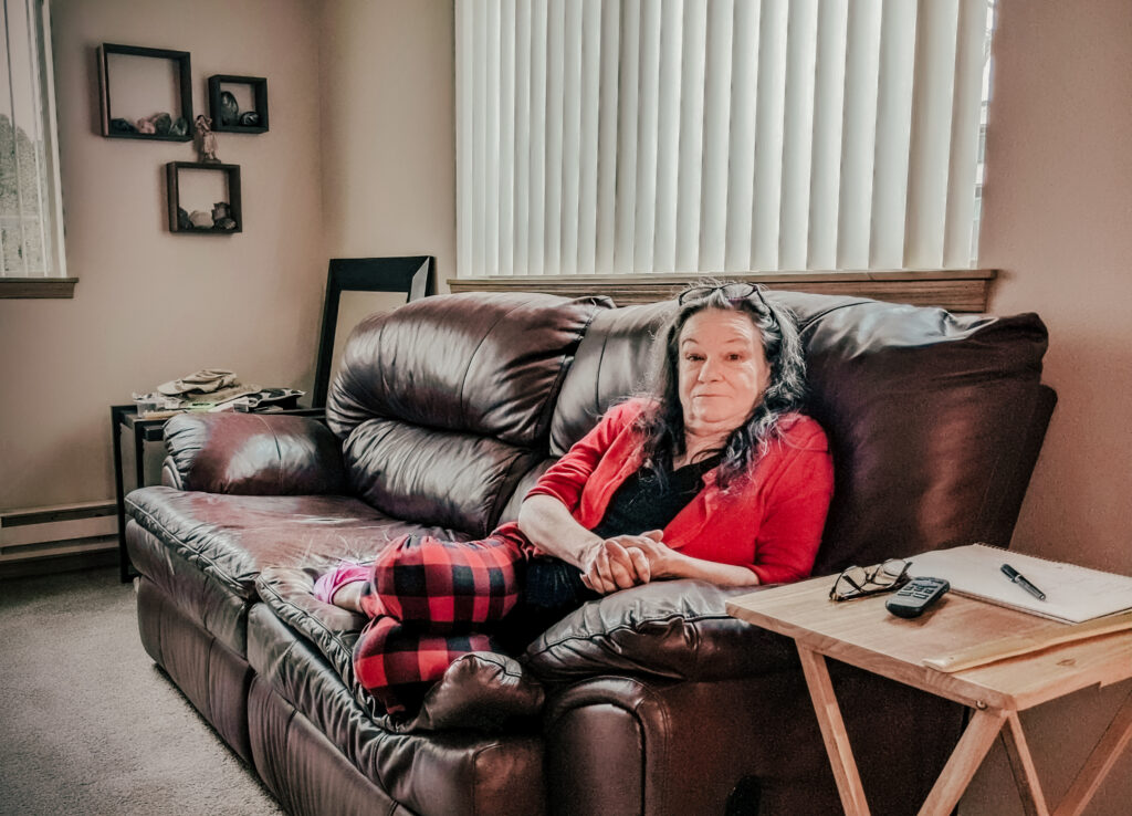 A woman wearing a red shirt and glasses resting on the top of her hair is sitting on a couch in an apartment living room.