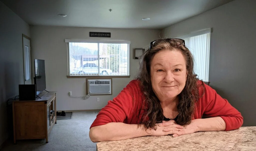 A woman wearing a red shirt and glasses resting on the top of her hair is sitting at a counter in a modest apartment kitchen and smiles at the camera.