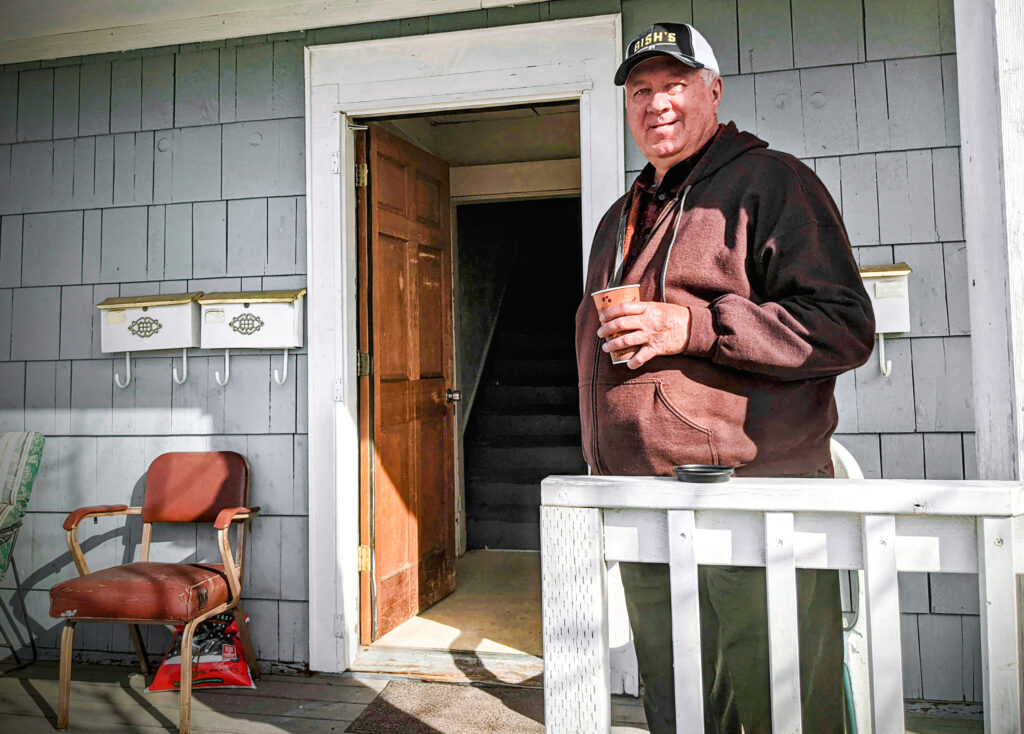 A man is standing on a porch, holding a coffee cup and looking and smiling at the camera.