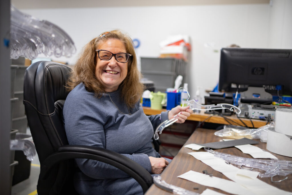 A woman is sitting in an office chair at a desk with empty hangers on it. She is smiling at the camera.