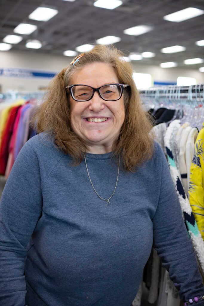A woman is standing in a thrift store, wearing a sweatshirt and smiling at the camera.