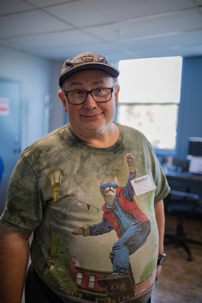 A man wearing a cap and glasses is standing in a room with computers and smiles at the camera.