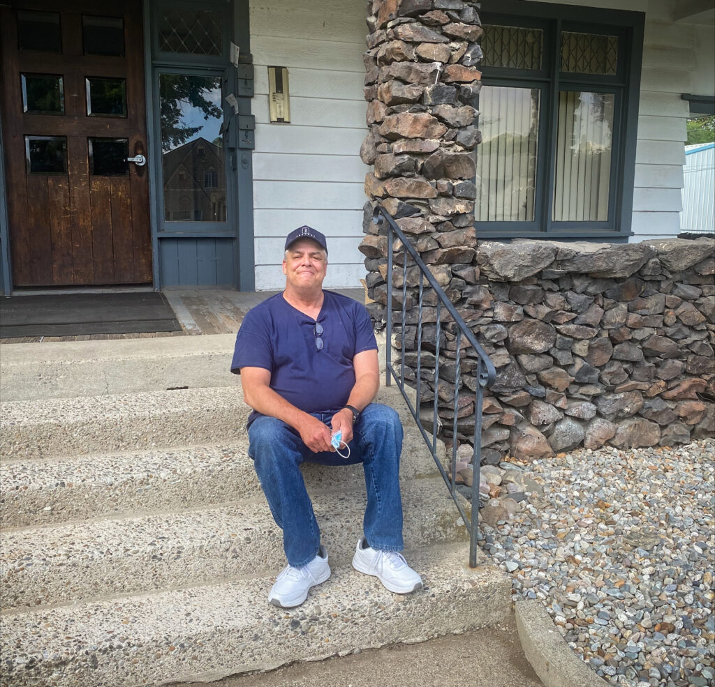 A man wearing jeans, a dark blue t-shirt and a ball cap is sitting on the front steps of an apartment building, smiling at the camera.