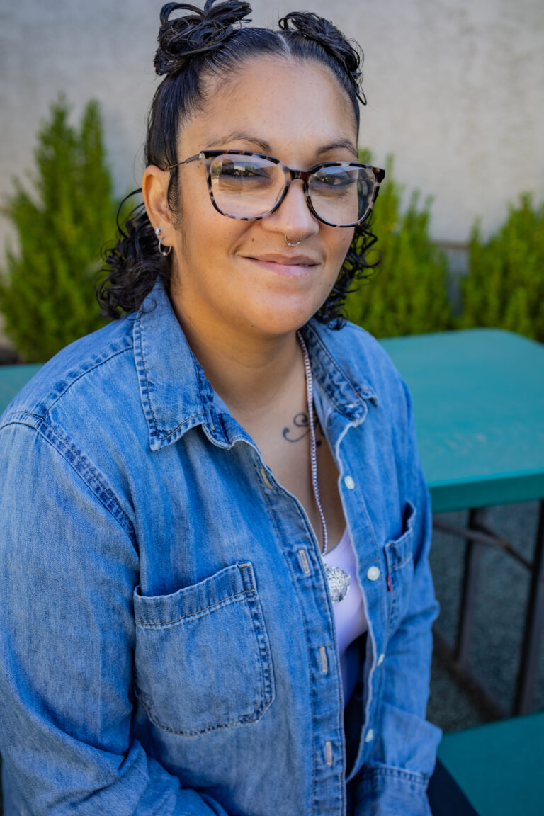 A woman wearing glasses and a denim button up shirt is sitting at a park bench and smiling at the camera.
