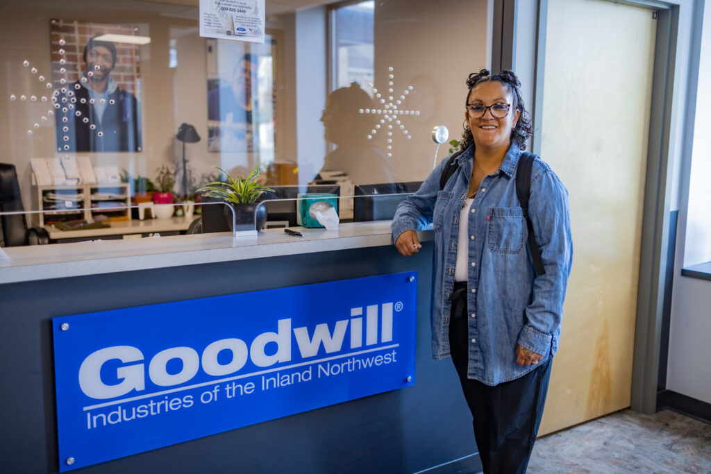 A woman wearing glasses and a denim button up shirt is standing at a counter in the lobby of a building. There is a blue sign below the counter that reads, "Goodwill Industries of the Inland Northwest"