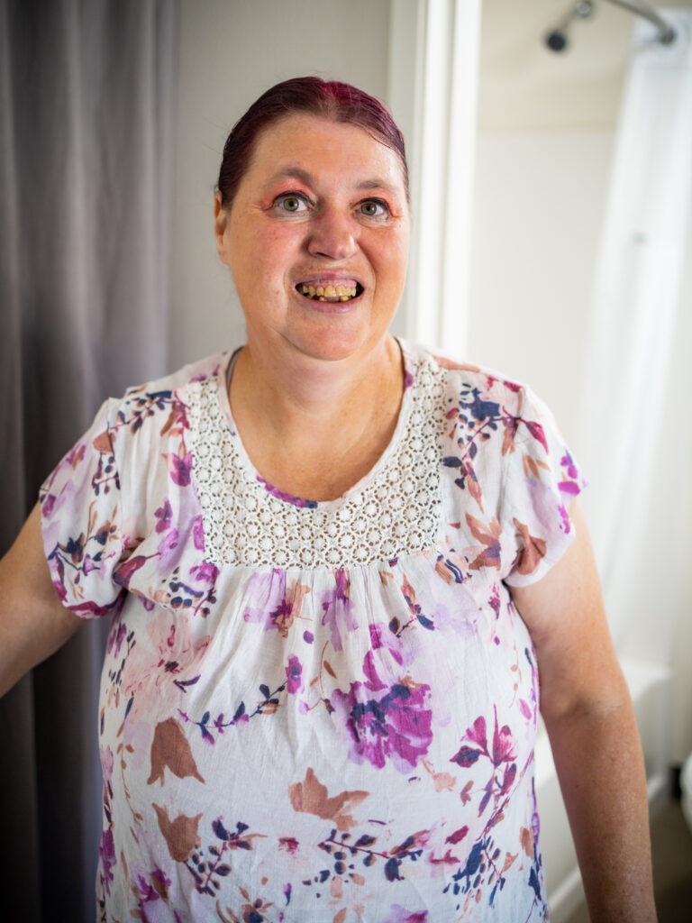 A woman wearing a floral patterned blouse is standing in a hotel room and smiling at the camera.