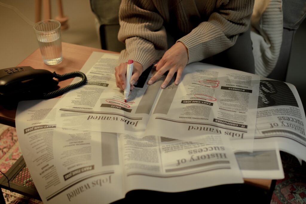 A woman holds a red marker and is circling job listings in a newspaper on a coffee table.