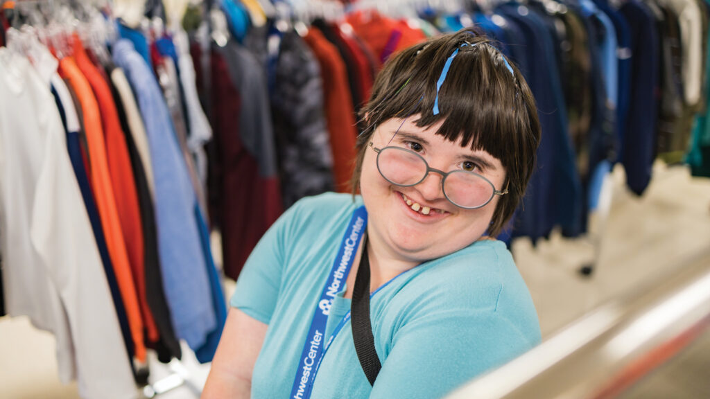 A girl wearing a light blue shirt and dark blue lanyard and glasses smiles at the camera. She stands in between racks of clothing.