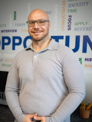 A man wearing glasses and a gray long sleeved and collared sweater is standing in front of a wall with words of affirmation and smiling.