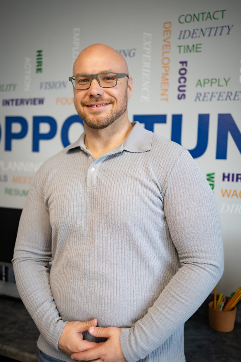 A man wearing glasses and a gray long sleeved and collared sweater is standing in front of a wall with words of affirmation and smiling.