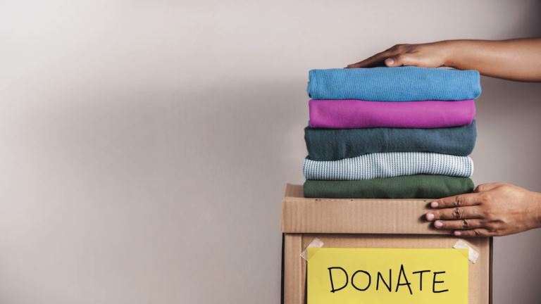 Two hands hold a stack of folded sweaters that sit atop a box with a handwritten sign that says "donate".