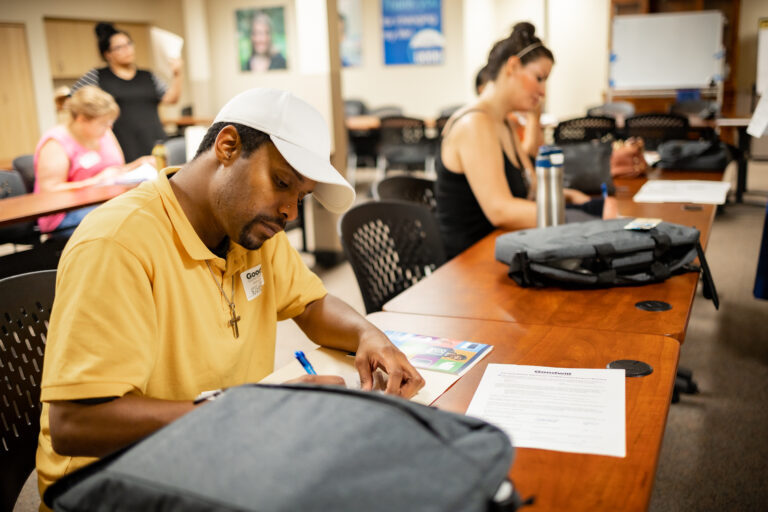 A man wearing a yellow collared shirt and a white ballcap is sitting at a long line of tables filling out paperwork. There is a laptop case next to him.