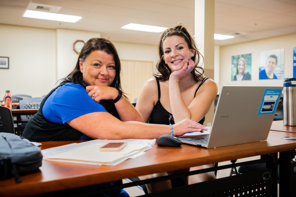 two women are at a desk with a laptop open in front of them. They are smiling at the camera.