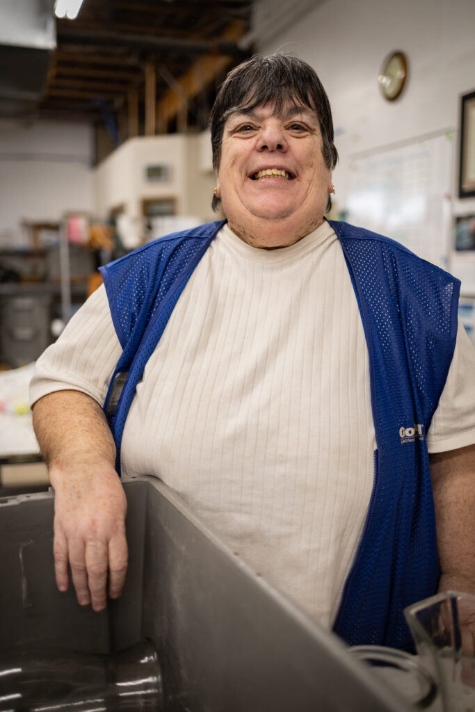 A woman wearing a white t-shirt and a dark blue vest is standing near a tub filled with wares and is smiling at the camera.