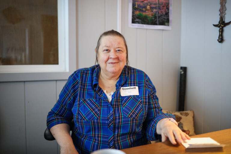 A woman wearing a plaid blue blouse is sitting at a table in an office and smiling at the camera.