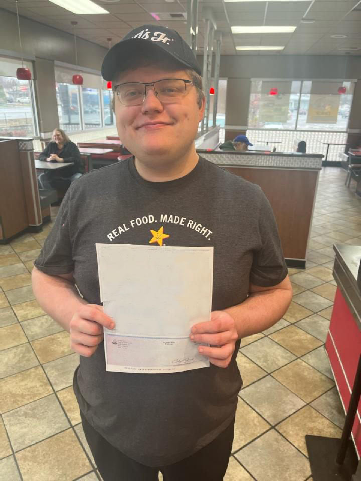 A man is holding a paycheck and standing in the lobby of a restaurant, smiling at the camera.