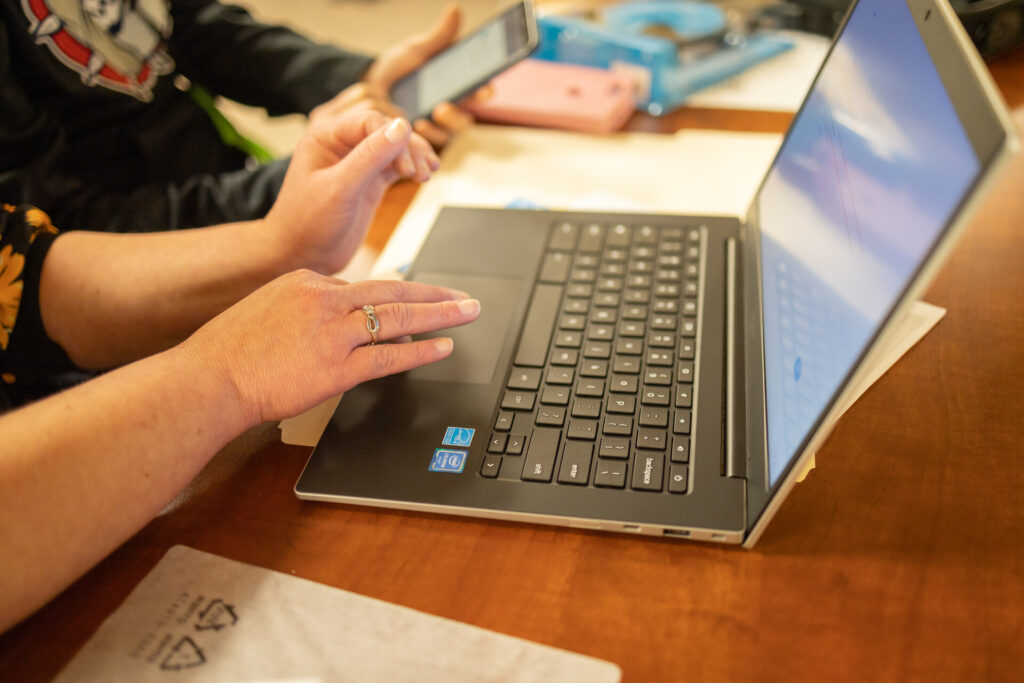 A woman's hands are mid-motion on a laptop keyboard. Another woman's hands hold a cell phone.