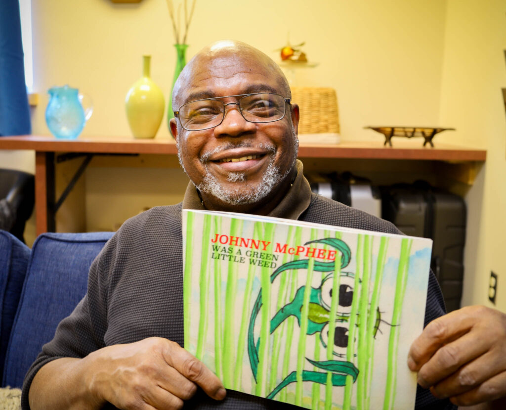 An ethnic man is sitting on a blue couch in an office, holding a children's book and smiling at the camera.