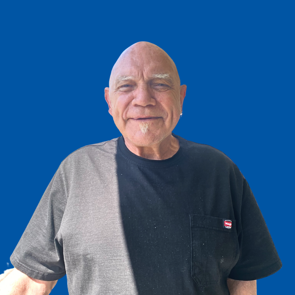 A man wearing a black t shirt is smiling at the camera. The background is a dark blue solid color.