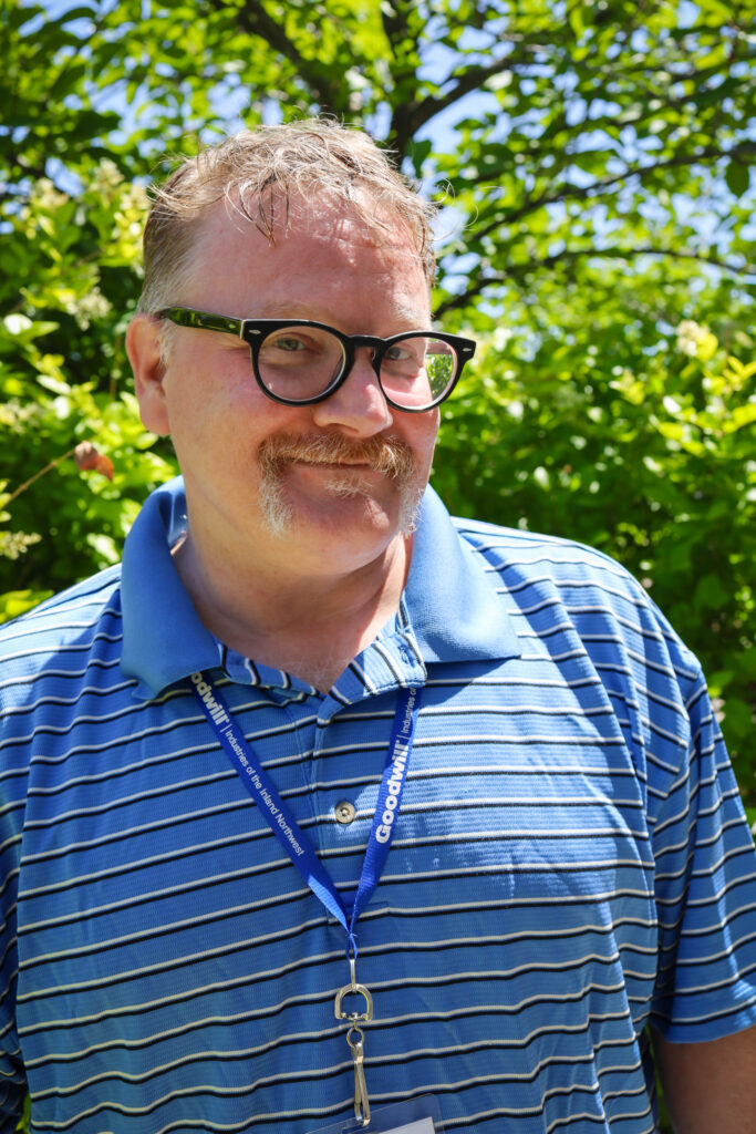 A man wearing glasses and a blue striped, collared shirt is standing in front of a large green bush and smiling at the camera.