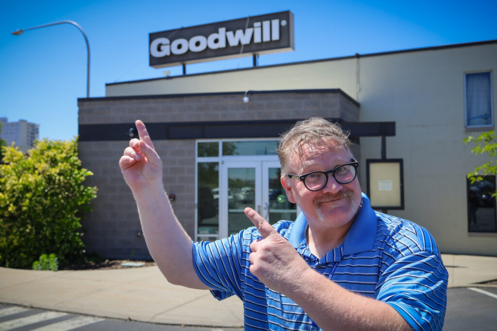 A man wearing glasses and a blue, striped, collared shirt is standing in front of a Goodwill building, smiling and pointing up at the Goodwill sign.
