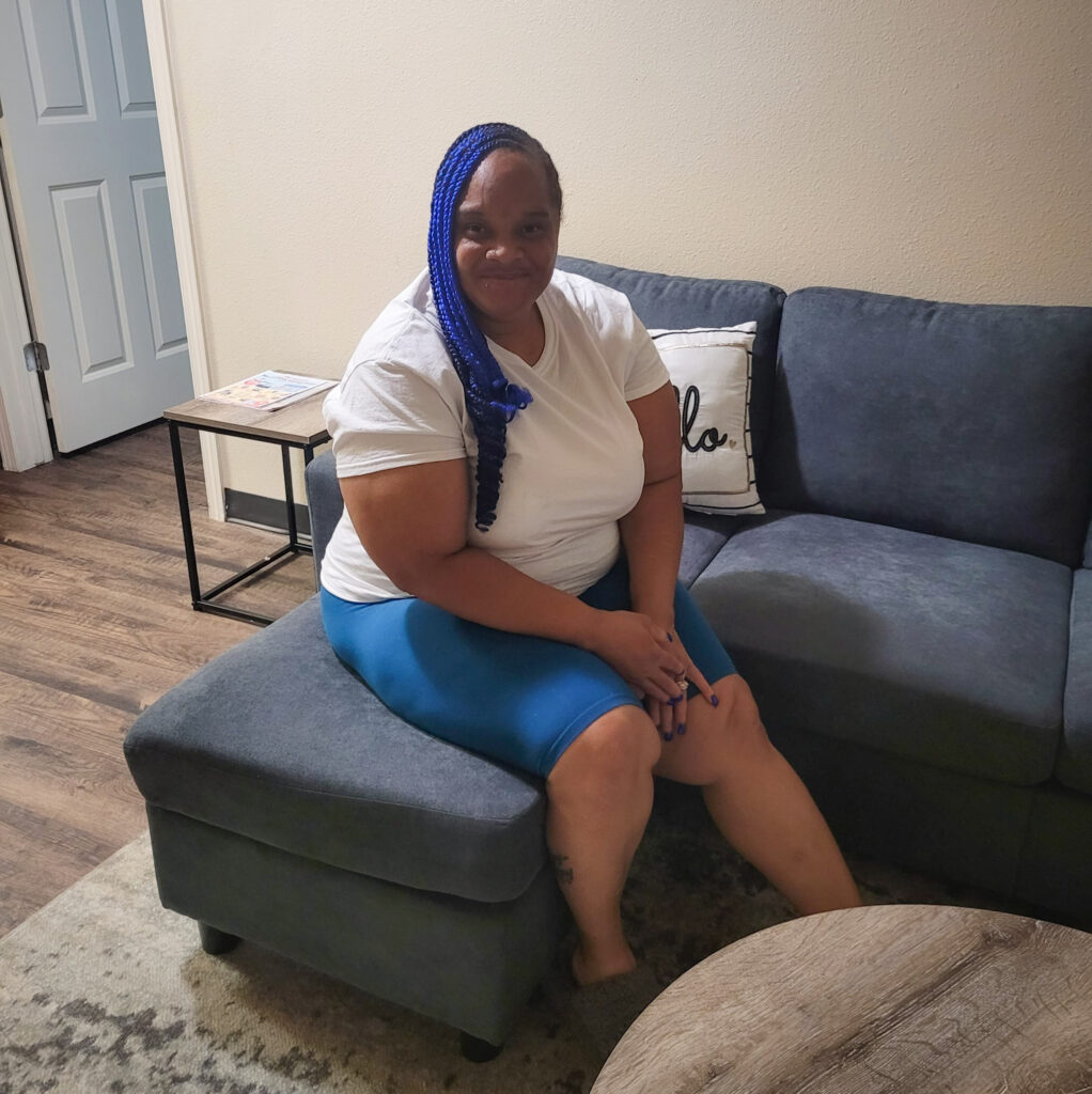 A woman with blue braids is sitting on a couch in a living room, smiling at the camera.