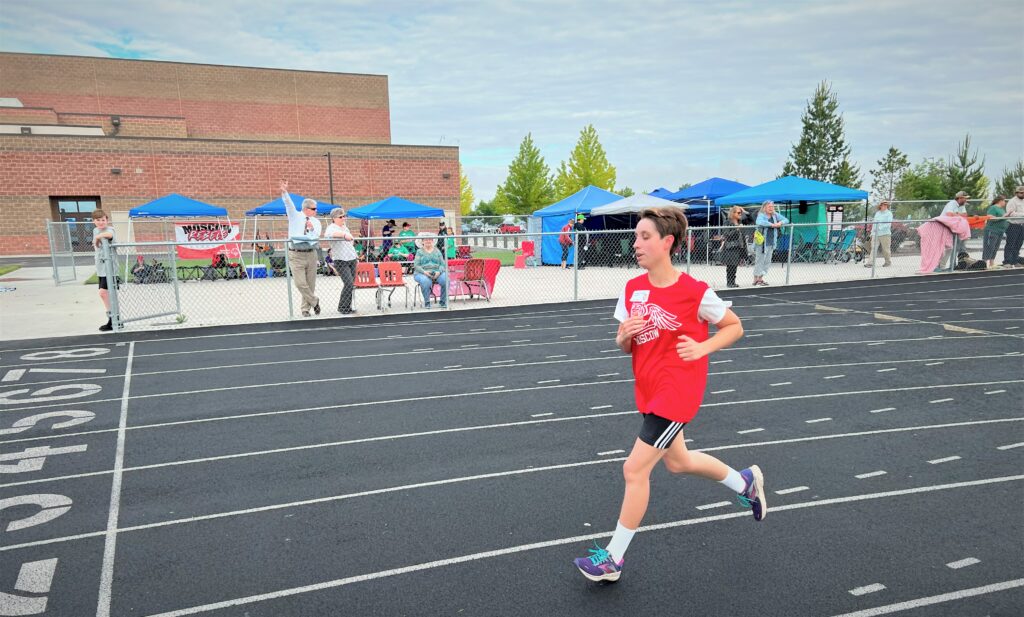 A woman wearing a red sports jersey is mid-run on a track.