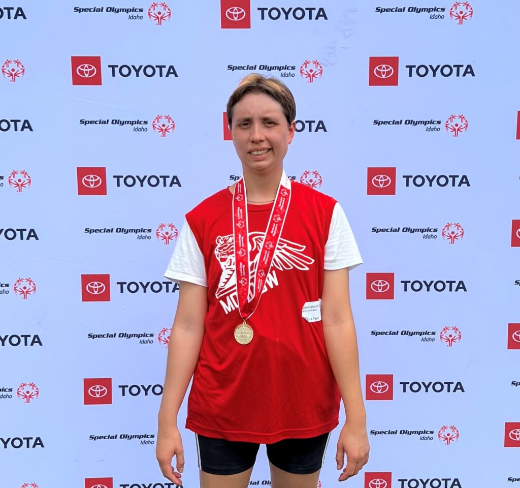 A woman wearing a red sports jersey is standing in front of a sponsored logo background, wearing a medal and smiling at the camera.