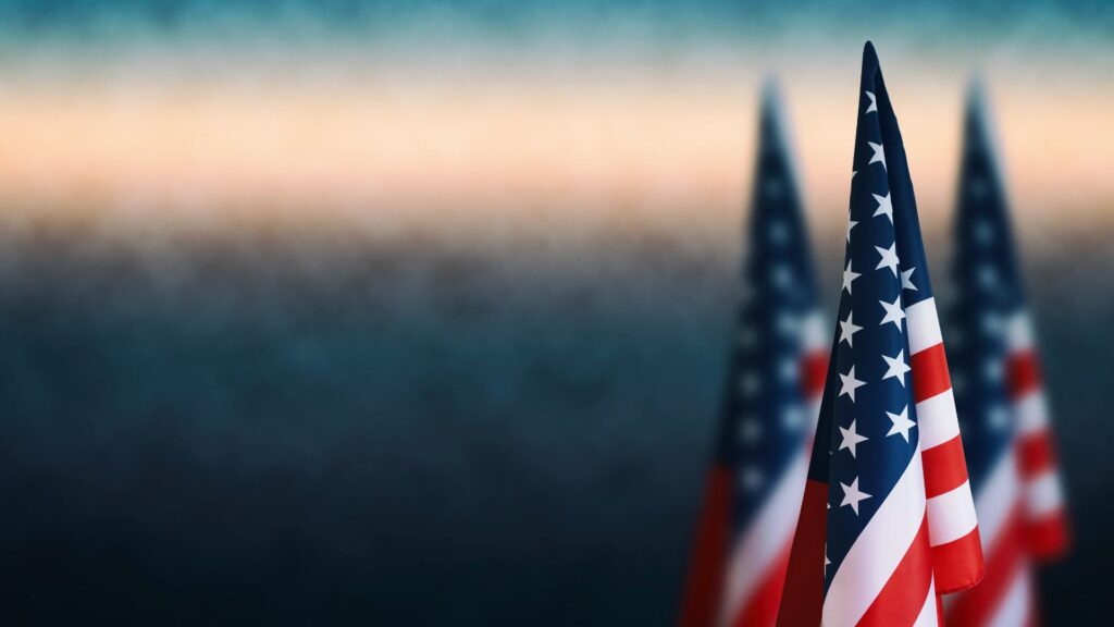 Three small American flags are in focus in the foreground with an out of focus gradient in the background. This image is for decoration.