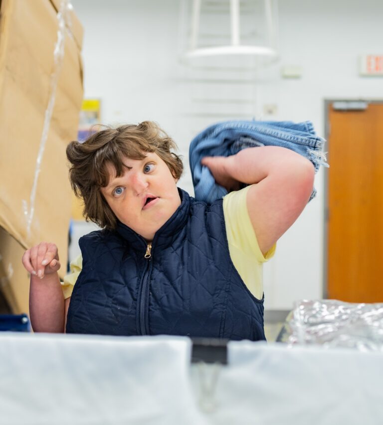 A woman wearing a yellow shirt and a zip-up vest is tossing textiles into a bin in a production area.