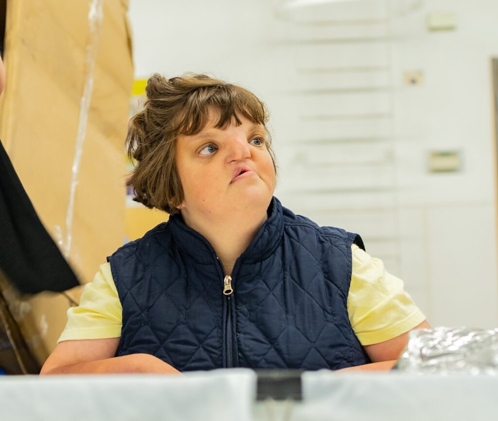 A woman wearing a yellow t-shirt and a zip-up vest is standing in a production area and looking off into the distance.