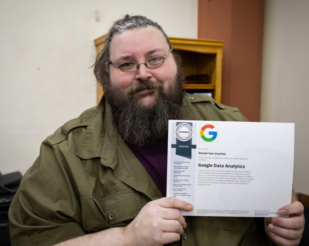 A long-haired, bearded man with glasses is holding up a Google Data Analytics certificate.