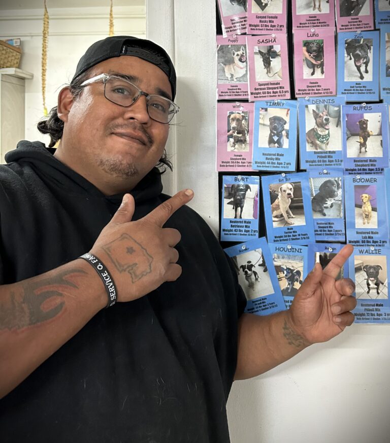 A man wearing a black sweater, backwards cap, and glasses is smiling and pointing to a wall with images of dogs available for adoption.