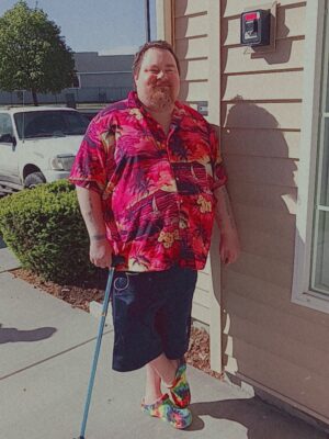 A man wearing a red Hawaiian shirt is leaning against a building wall and smiling at the camera.