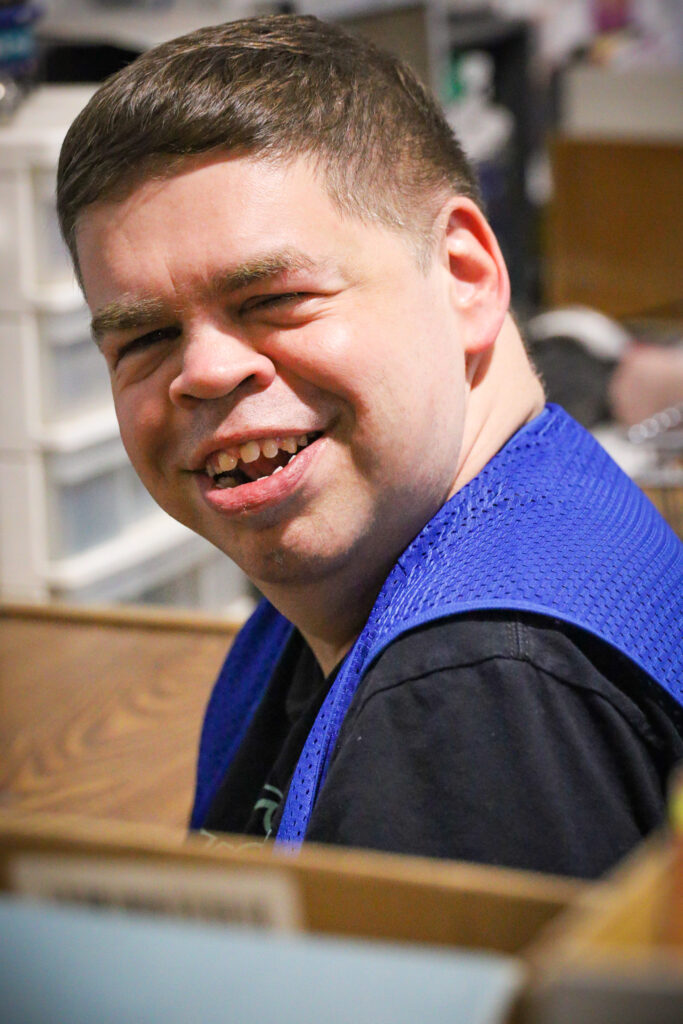 A close up of a man wearing a blue work vest and smiling at the camera.