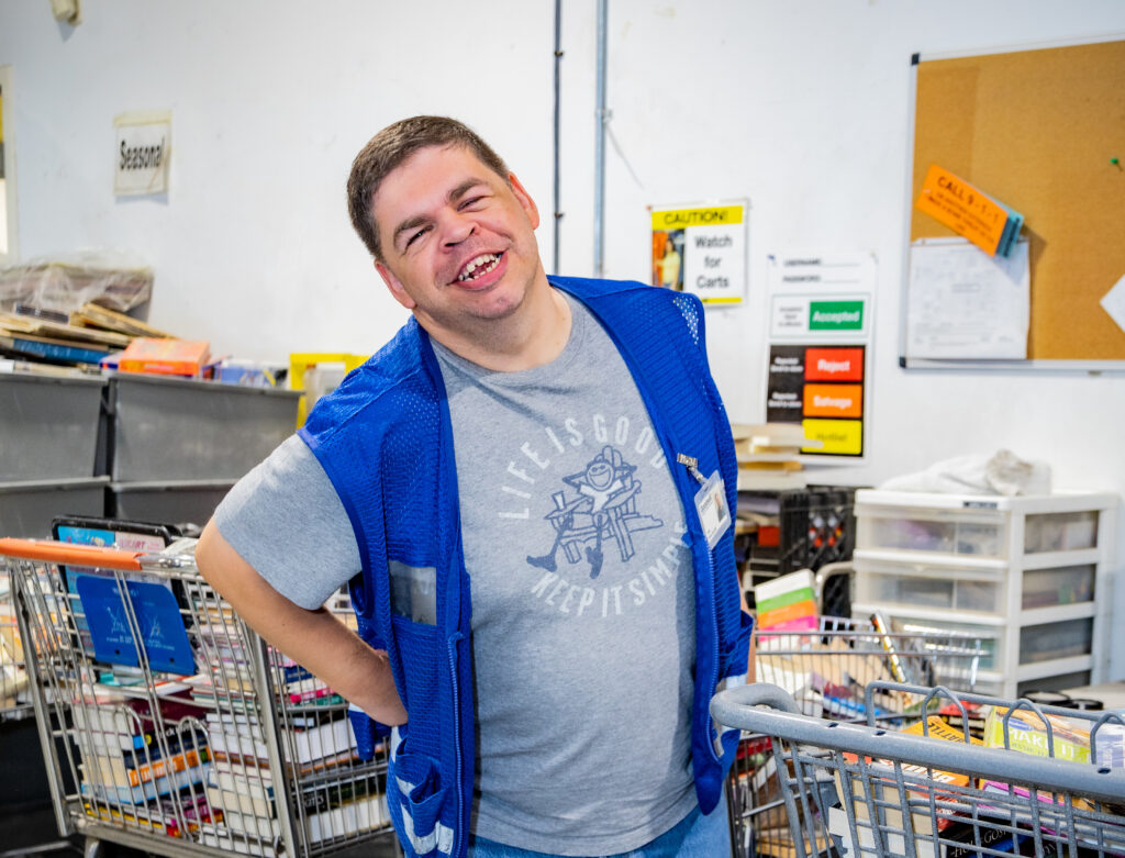 A man wearing a blue work vest and standing in a warehouse is posing and smiling for the camera.