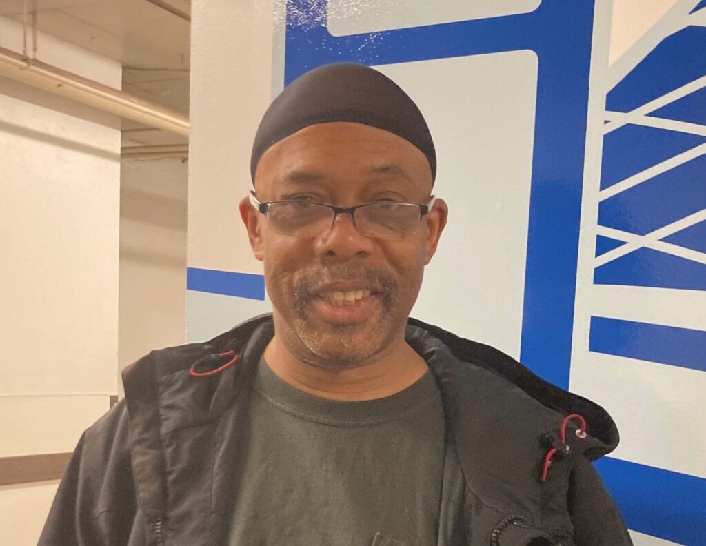 A man wearing a cap and glasses is standing in a facility and smiling at the camera.