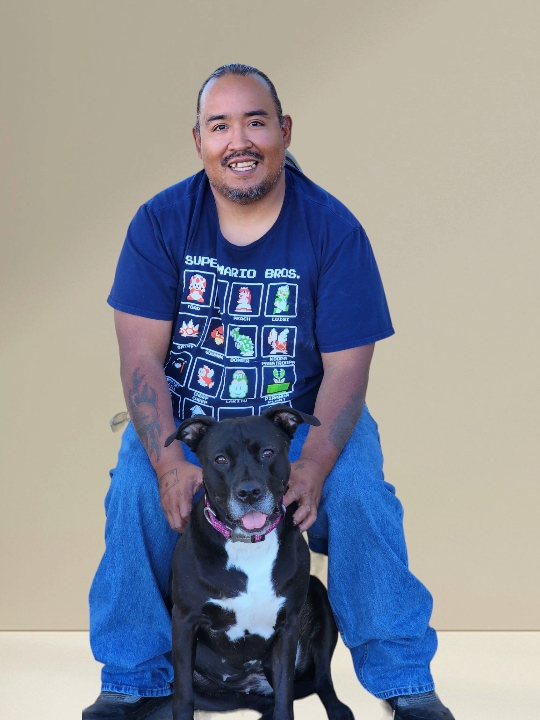 A man is standing over a black dog, petting it and smiling at the camera.